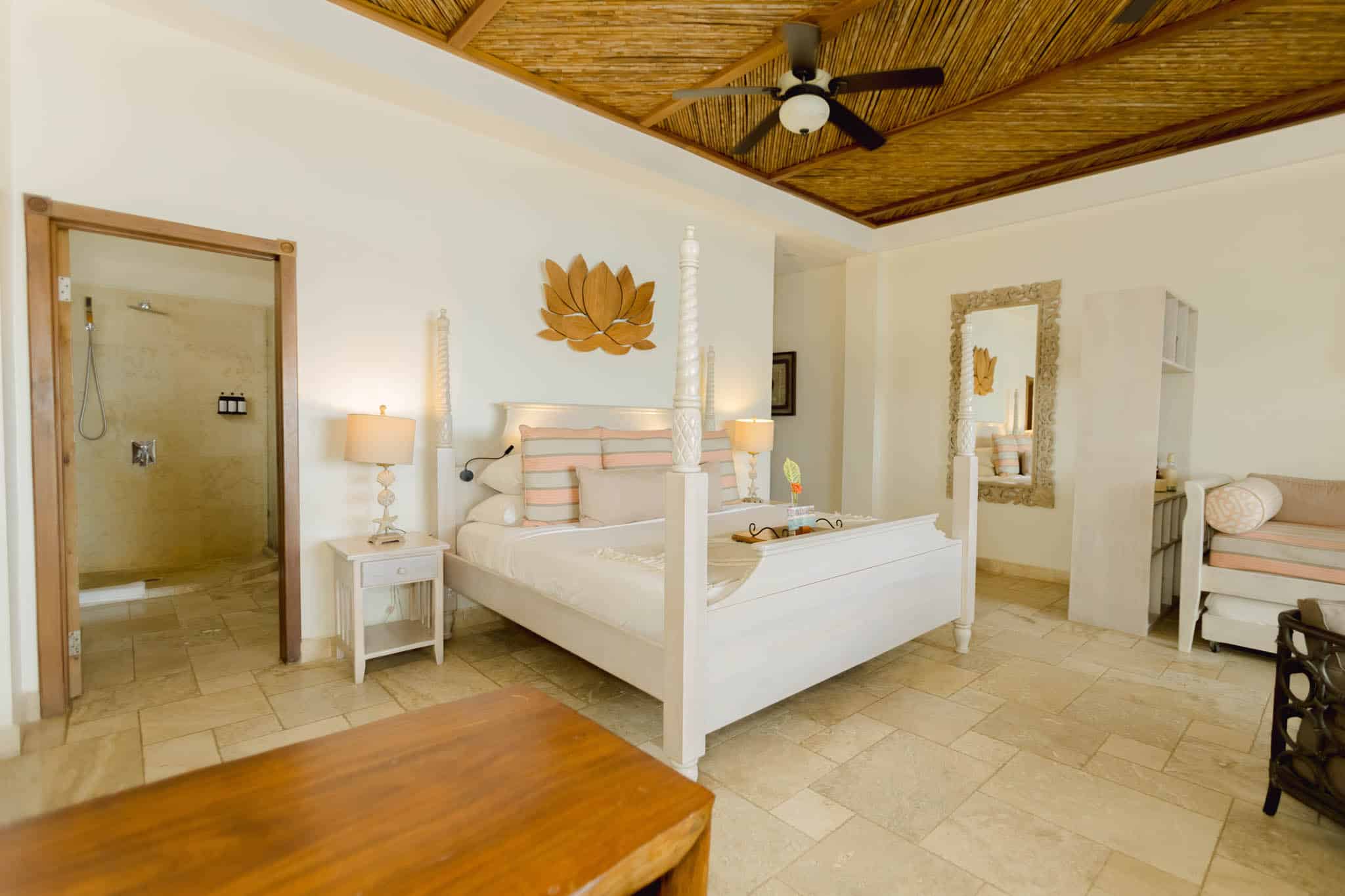 A bright, all-white bedroom in a Beachfront Spa Room at Almond Beach featuring a white bedframe, a decorative wooden structure hanging above the bed, and a ceiling fan.