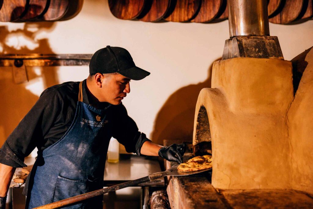 A chef in a black cap and denim apron carefully placing a small pizza into a traditional wood-fired clay oven with a metal peel at Don Tonito's, Jaguar Reef.